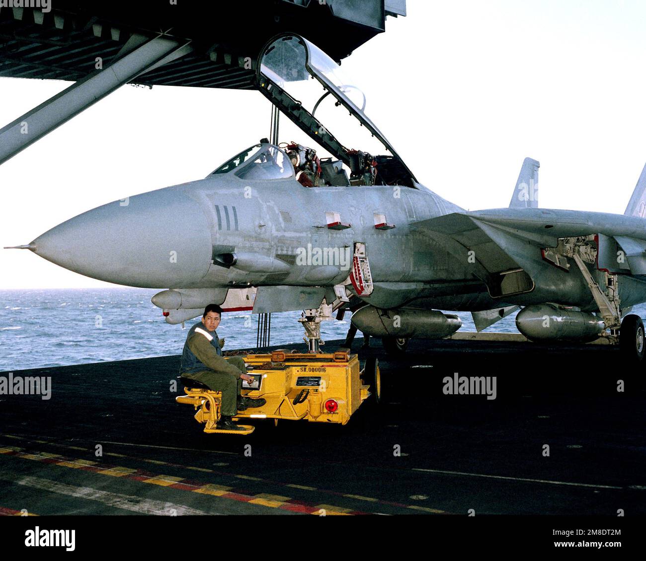 Ein Besatzungsmitglied des Cockpits steht mit einem SD-1D bereit, der neben einem Fighter Squadron 14 (VF-14) F-14A Tomcat-Flugzeug einen Dolly beobachtet, während das Flugzeug von einem abgesenkten Aufzug an Bord des Flugzeugträgers USS JOHN F. KENNEDY (CV 67) zum Abschleppen vorbereitet ist. Basis: USS John F. Kennedy (CV 67) Stockfoto