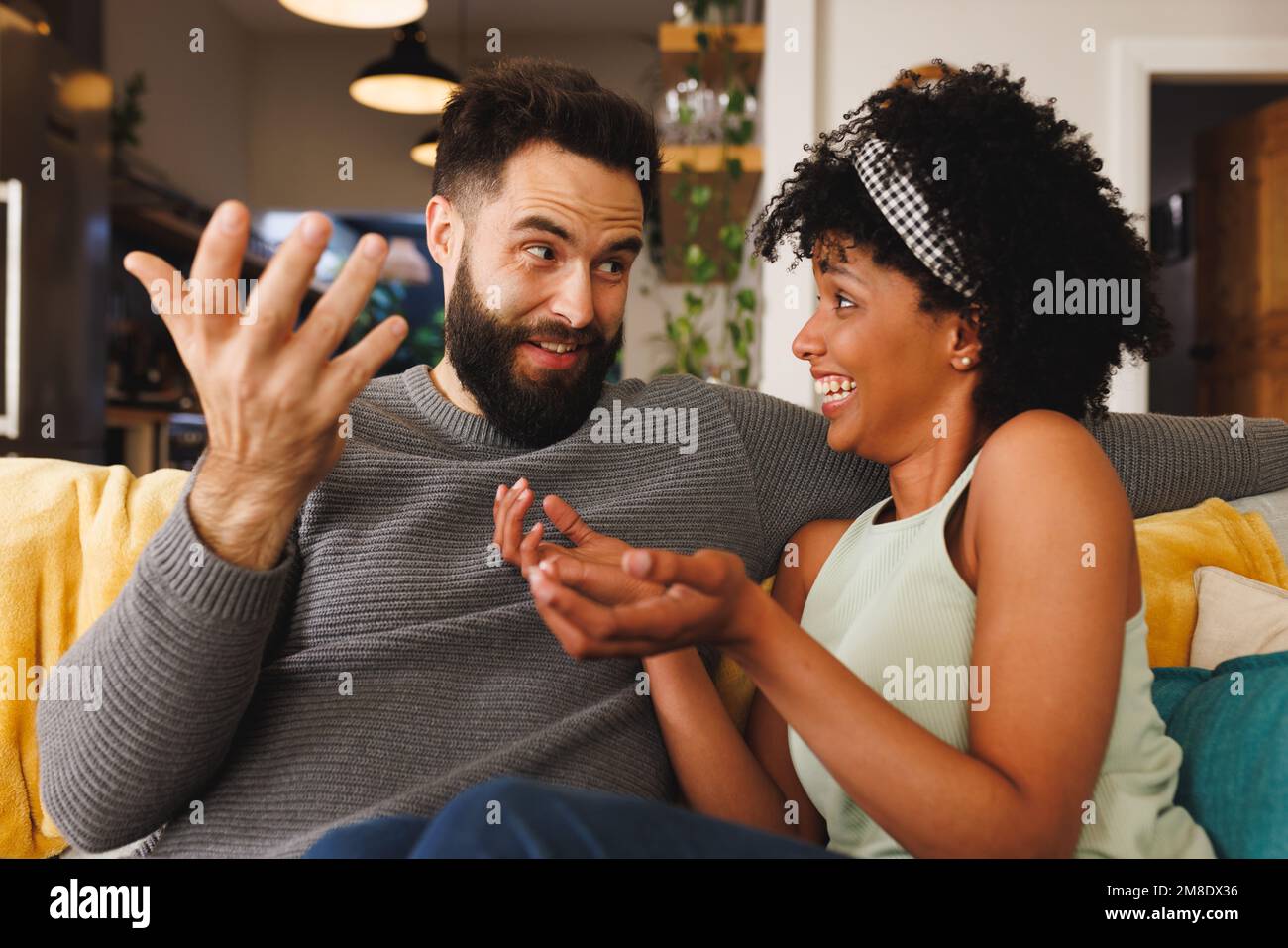 Glückliches birassisches junges Paar, das sich ansieht und redet, während es auf dem Sofa im Wohnzimmer sitzt Stockfoto