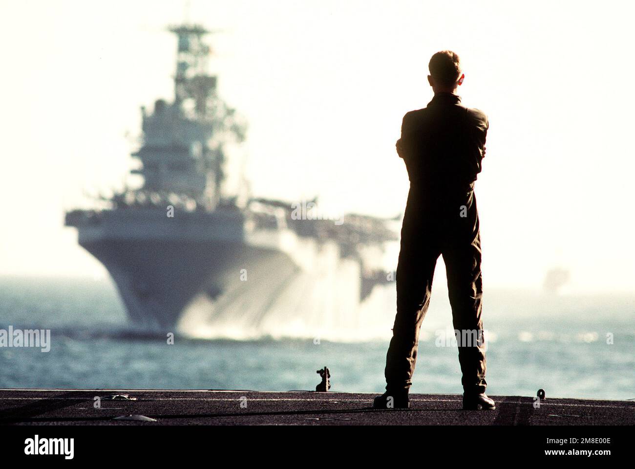 Ein Marine an Bord der Amphibien-Anlegestelle USS RALEIGH (LPD-1) beobachtet, wie das Amphibienschiff USS NASSAU (LHA-4) aus DEM Steuerbordviertel von RALEIGH dampft. Die Schiffe und ihre Marines sind auf See, um Operation Desert Shield zu unterstützen. Betrifft Operation/Serie: WÜSTENSCHILDLAND: Nördliches Arabisches Meer Stockfoto