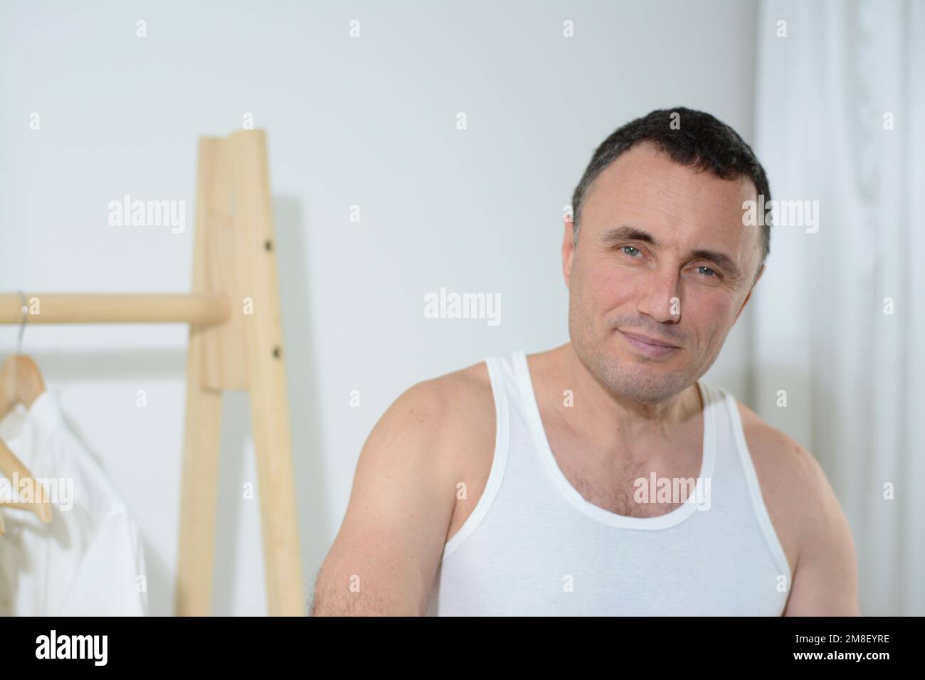 Der gutaussehende Mann steht und schaut mit blauen Augen in den Rahmen. Er hat einen müden Blick oder er wachte morgens im Kleiderbügel auf, mit Hemden. Er trägt ein T-Shirt und macht sich auf die Arbeit Stockfoto