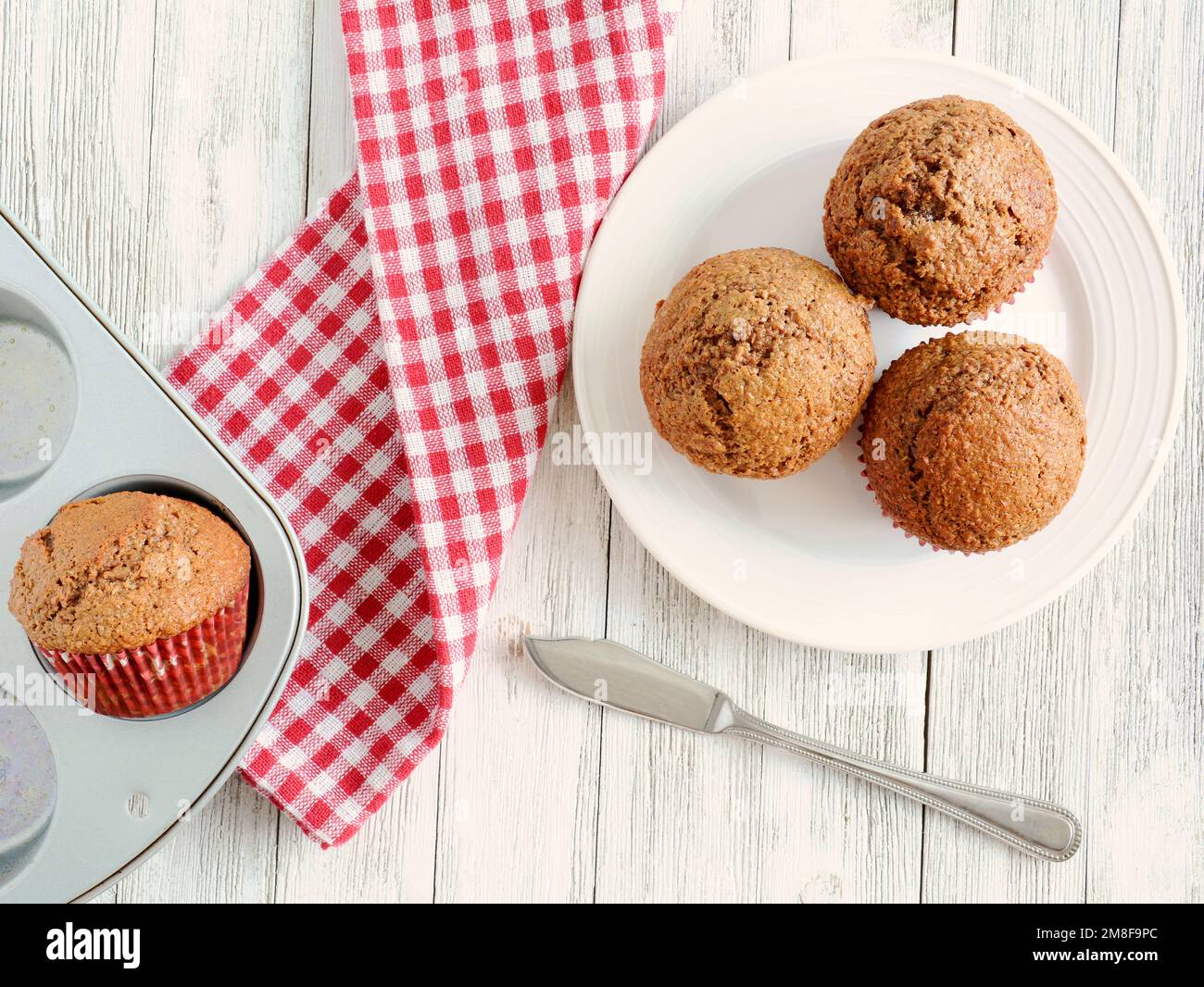 Frisch gebackene Kleie-Muffins auf weißem Teller mit rot kariertem Geschirrtuch und Muffin-Pfanne auf rustikalem weißem Holz in flacher Lage. Stockfoto