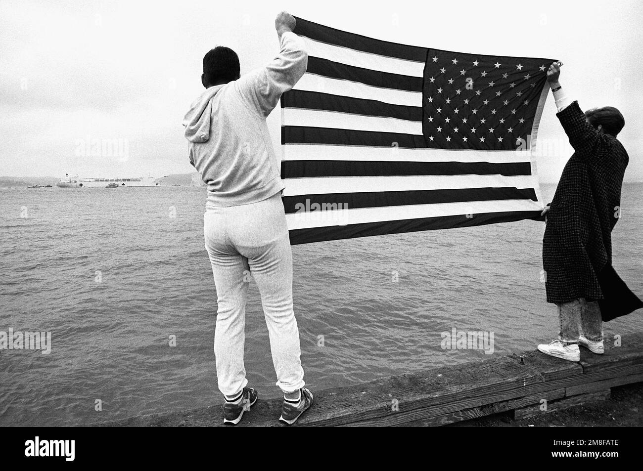 Die Angehörigen eines Besatzungsmitglieds an Bord des Krankenhausschiffs USNS MERCY (T-AH-19) zeigen eine amerikanische Flagge auf dem Pier, während das Schiff in Sicht kommt. Die GNADE kehrt nach San Francisco zurück, nachdem sie während der Operation Desert Storm im Persischen Golf eingesetzt wurde. Betreff Operation/Serie: WÜSTENSTURMBASIS: San Francisco Bay Bundesstaat: Kalifornien (CA) Land: Vereinigte Staaten von Amerika (USA) Stockfoto