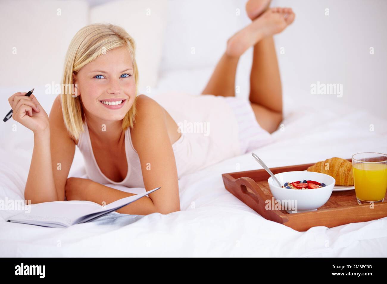 Eine To-Do-Liste für den Tag aufschreiben. Blonde Frau, die auf ihrem Bett liegt und in ihr Tagebuch schreibt, mit ihrem Frühstück neben ihr - Porträt. Stockfoto