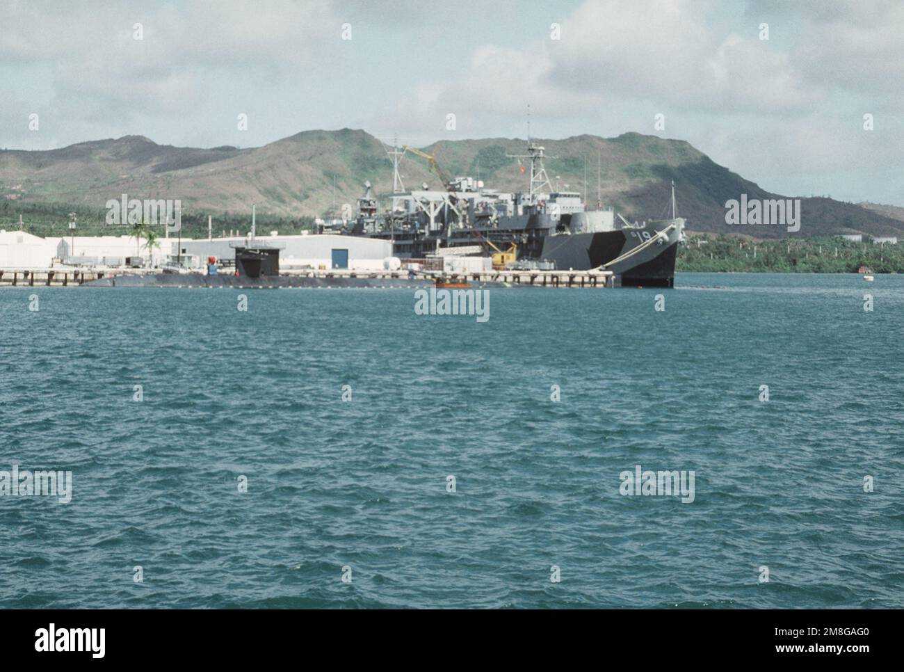 Der U-Boot-Tender USS PROTEUS (AS-19) und ein nicht identifizierter U-Boot-Stand, der am Pier am Marinestützpunkt festgemacht ist. Das TARNMUSTER VON PROTEUS aus dem Zweiten Weltkrieg wurde für den Besuch des Schiffs in Australien anlässlich der Zeremonien anlässlich des 50. Jahrestages der Schlacht um die KORALLENSEE hinzugefügt. Basis: Marinestützpunkt Staat: Guam (GU) Land: Nördliche Marianen (MNP) Stockfoto