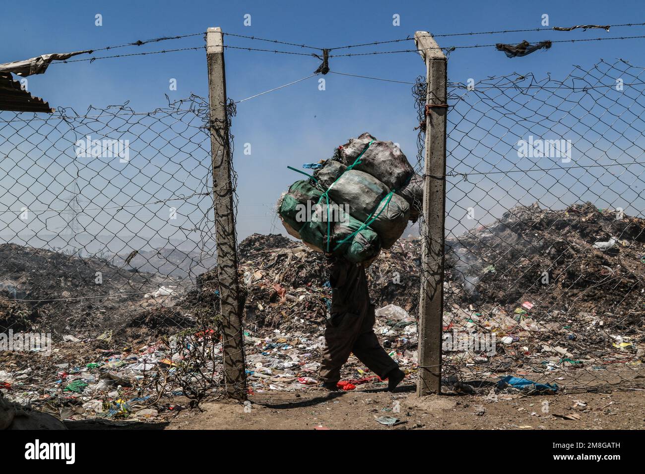 Ein Abfallsammler transportiert Müllsäcke mit Abfall zum Recycling neben dem brennenden Müll auf dem Hauptdepot von Nakuru. Da Abfälle selten getrennt und stark mit allen Arten von Abfall vermischt werden, kann das Verbrennen potenziell giftige Dämpfe erzeugen, die ein Problem für die öffentliche Gesundheit darstellen und auch zum Klimawandel beitragen können. Gemeinden, die in der Nähe von Mülldeponien arbeiten und leben, wo Müll offen verbrannt wird, sind dem Risiko von Atemwegserkrankungen und Krebs ausgesetzt. Frauen, die diesen Dämpfen ausgesetzt sind, können unter reproduktiven Gesundheitsproblemen leiden. Stockfoto