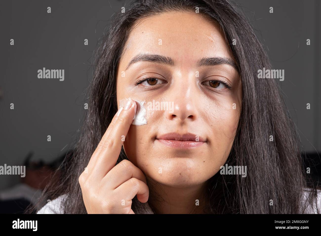 Kaukasische Brünette, die in die Kamera schaut und Reinigungscreme auf ihre Wange aufträgt. Nahaufnahme des Kopfaufnahmen-Porträts einer Hautbehandlungskonzeption einer jungen Frau. Stockfoto