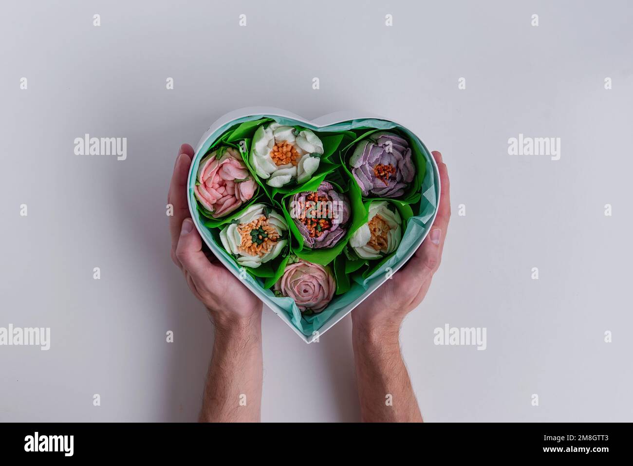 Auf einem weißen, isolierten Hintergrund halten männliche Hände eine herzförmige Geschenkbox mit süßen Marshmallow-Blumen. Natürlich handgefertigt. Nachtisch ohne Zucker Stockfoto