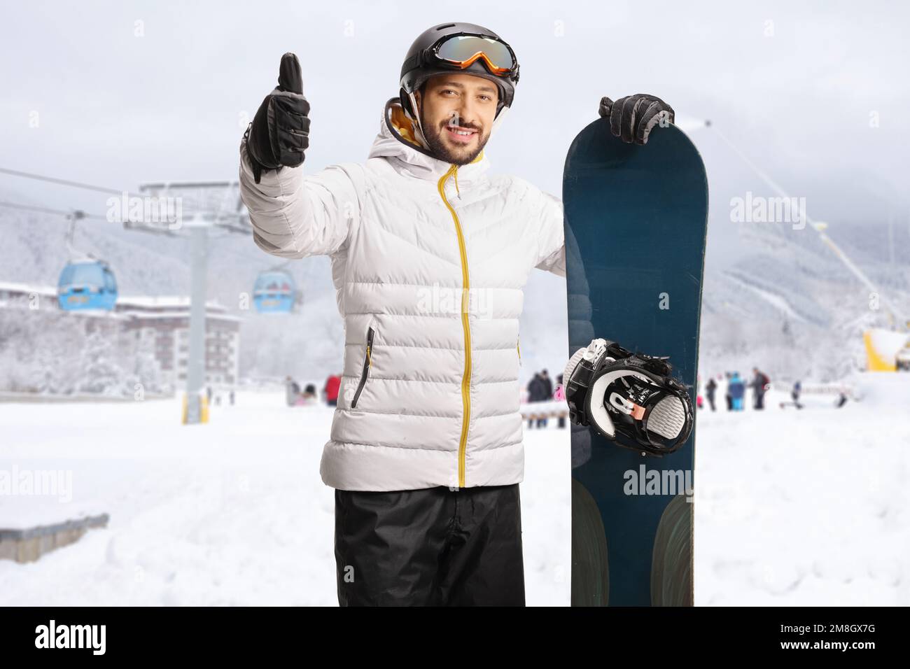 Ein Typ mit einem Snowboard, der in einem Skigebiet steht und Daumen nach oben zeigt Stockfoto