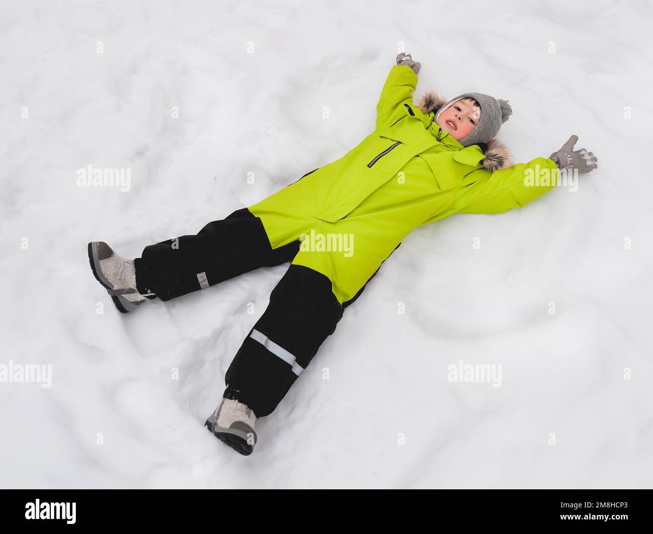 Ein lächelnder Junge im grünen Overall macht Schneeengel auf Schnee. Fröhliches Kind, das bei Schneewetter im Freien spielt. Draufsicht auf ein glückliches Kind Stockfoto