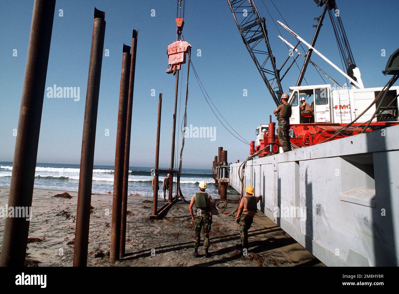 USA Army Corps of Engineers von der 497. Engineering Company, verwenden Stahlschablone, um sechs Zoll große Pfähle zu verteilen, die den Rahmen des neuen Grenzzauns bilden, der die Vereinigten Staaten und Mexiko trennt. Sie verwenden dann einen zwei-Tonnen-Hammer, der eine Kraft von 7.000 Pfund hat, um die Pfähle einzutreiben. Der Kran hielt die Hammerablage auf dem erhöhten Damm, der von den Navy Seabees des Amphibious Construction Battalion One (ACB-1) als Teil der Joint Task Force Six (JTF6) gebaut wurde. Die Grenzpolizei wird Stahlmatten am Zaun anbringen. Basis: Border Field St Park San Diego Staat: California (CA) Cou Stockfoto
