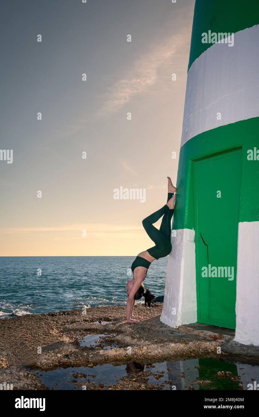 Eine sportliche Frau, die einen Handstand an der Wand eines Leuchtturms am Meer hält. Stockfoto