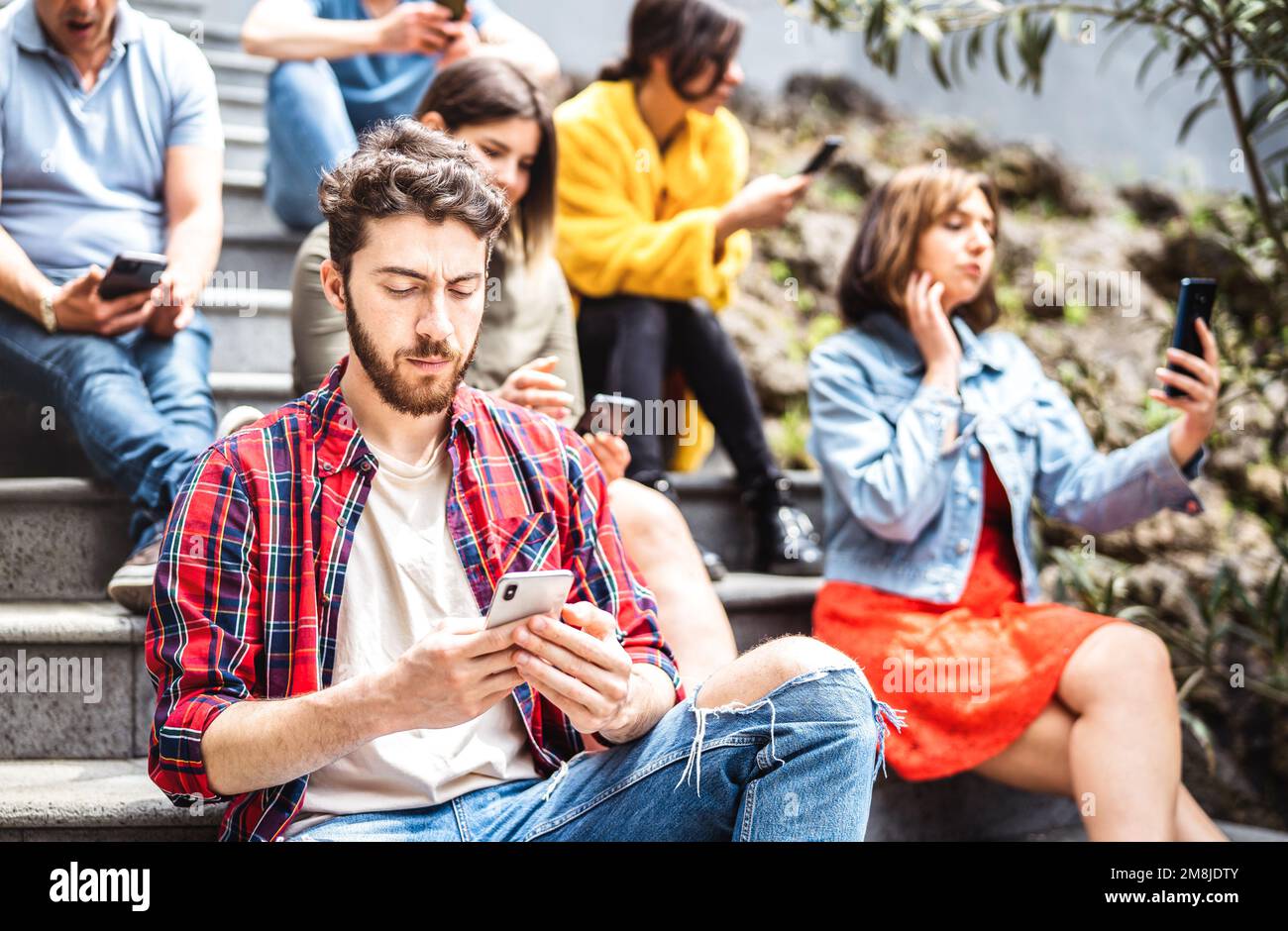 Gelangweilte Gruppe, die ein Smartphone benutzt und auf dem Hof des städtischen Colleges sitzt - Junge Freunde, die von Smartphone-Geräten abhängig sind - Technologiekonzept WIT Stockfoto