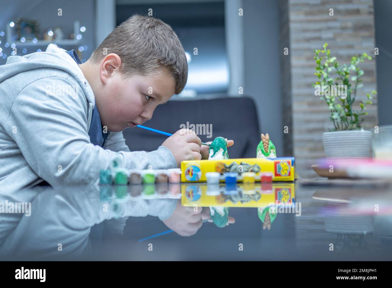 Ein Junge färbt eine Dinosaurierfigur. Er ist sehr konzentriert und ernst. Er hält einen Pinsel und trägt vorsichtig Farbe auf die Figur auf Stockfoto