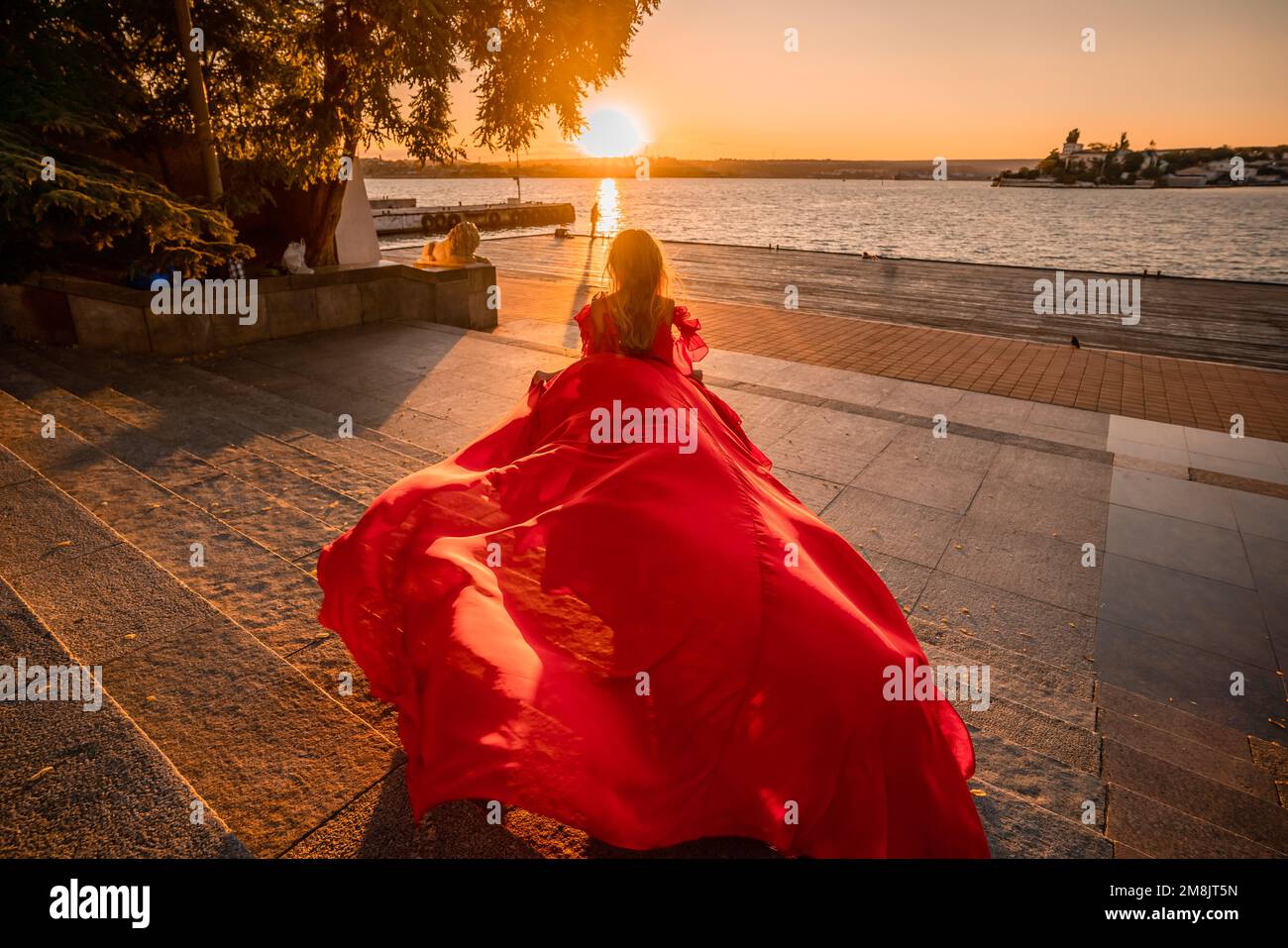 Rotes Kleid bei Sonnenaufgang. Eine Frau in einem langen roten Kleid vor dem Hintergrund des Sonnenaufgangs, helles goldenes Licht der Sonnenstrahlen. Der Begriff der Weiblichkeit Stockfoto