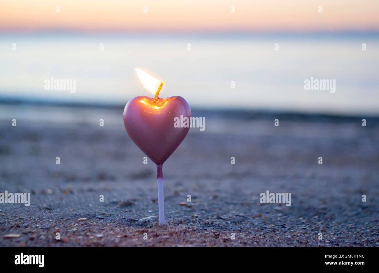 Herzförmige Kerze, die bei Sonnenaufgang auf dem Sandstrand in der Nähe von Meereswellen brennt. Brennende Kerze in Form eines Herzens im Sand vor dem Hintergrund der aufgehenden Sonne. Romantische romantische Stimmung. St. Valentinstag. Stockfoto