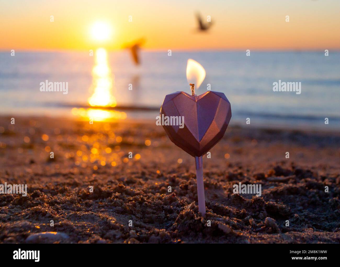 Herzförmige Kerze, die bei Sonnenaufgang auf dem Sandstrand in der Nähe von Meereswellen brennt. Brennende Kerze in Form eines Herzens im Sand vor dem Hintergrund der aufgehenden Sonne. Romantische romantische Stimmung. St. Valentinstag. Stockfoto