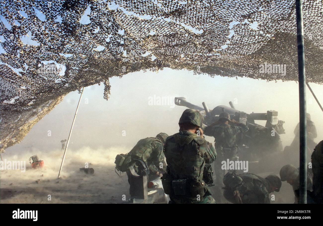 Rückansicht links, Kamera unter Tarnnetz. Ein M198 155-mm-Howitzer. Geschützführer im Vordergrund, zwei Crewmitglieder bücken sich, um ein 155mm-Projektil aufzunehmen. Land: Unbekannt Stockfoto