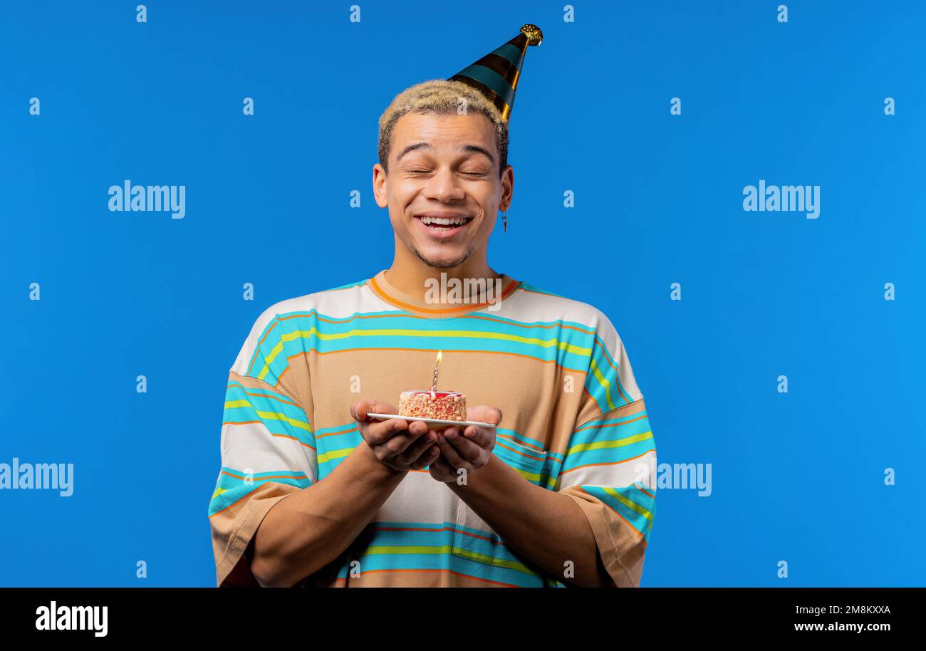 Alles gute zum Geburtstag, Mann, der sich was wünscht - Kerze auf Kuchen blasen. Ein lockiger Kerl lacht, feiert seinen Jahrestag. Junger Teenager mit blauem Hintergrund. Stockfoto