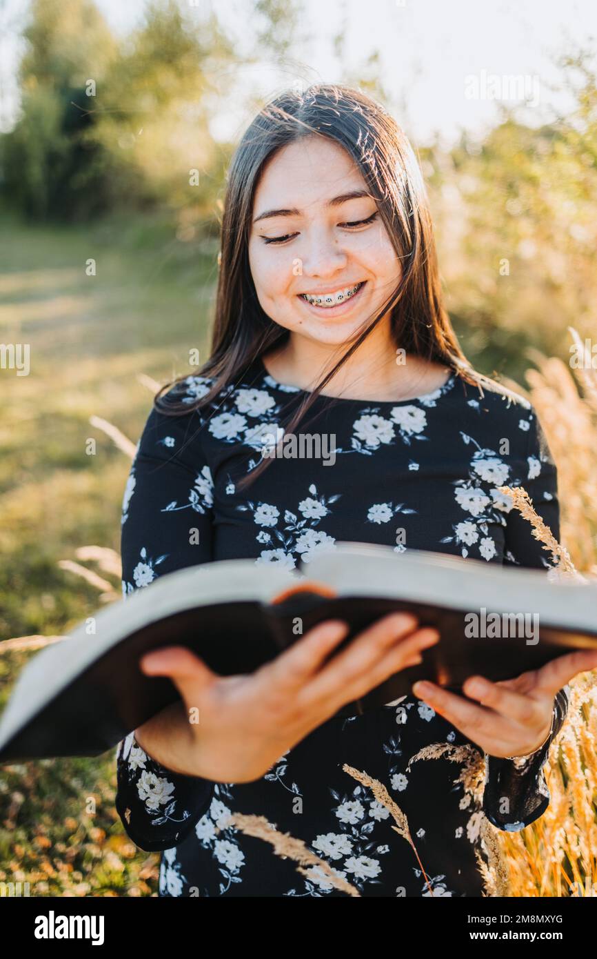 Lächelndes junges religiöses Mädchen, das ihre bibel liest, draußen auf dem Feld bei Sonnenuntergang. Spirituelle Wiederbelebung. Stockfoto