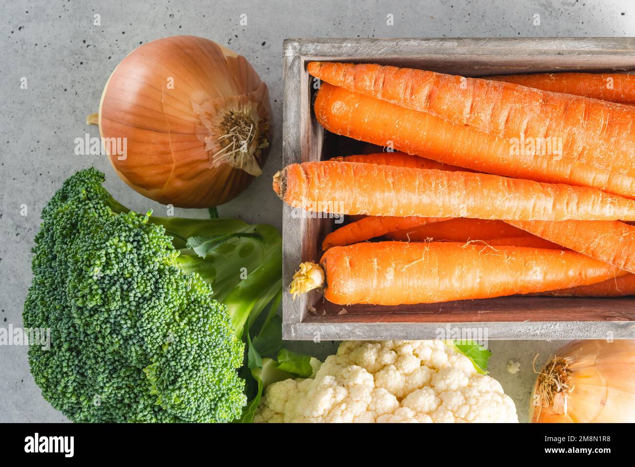 Karotten in einer Holzkiste, Brokkoli, Blumenkohl und Zwiebeln – Nahaufnahme auf grauem Steinhintergrund, flach liegend mit Kopierbereich Stockfoto