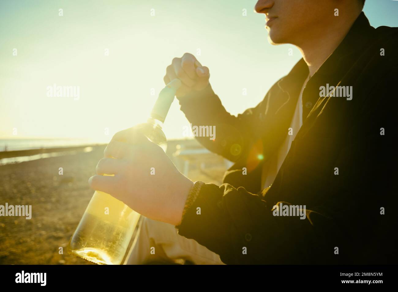 Ein Mann am Ufer des Meeres oder Ozeans öffnet eine Flasche Weißwein. Ein Date am Strand, Alkohol auf der Straße vor dem Hintergrund des Sonnenuntergangs Stockfoto