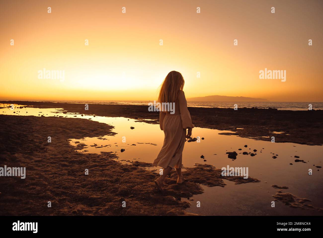 Ein wunderschönes Mädchen in einem Kleid vor dem Hintergrund des Sonnenuntergangs im Hintergrund des Meeres, eine Frau mit langen Haaren im Urlaub. Unglaubliche Natur, das oc Stockfoto