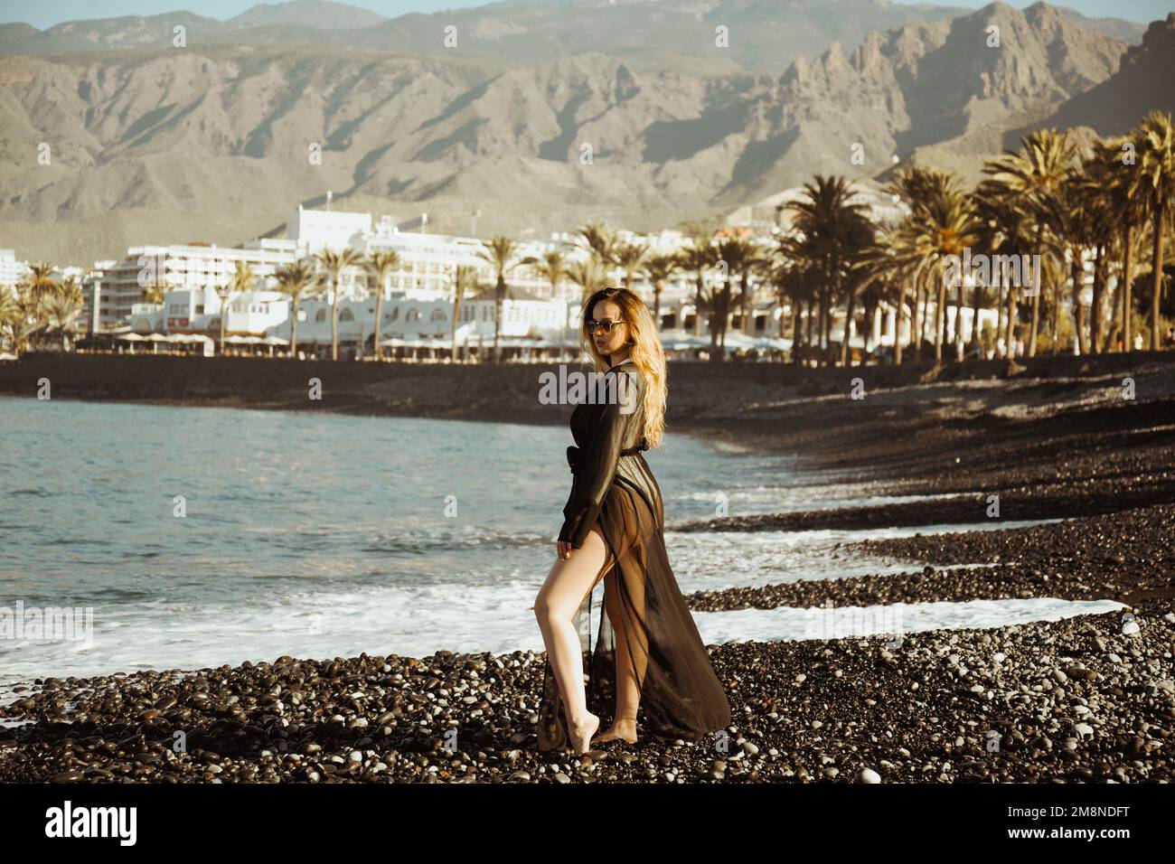 Ein Mädchen geht an einem Steinstrand in einem langen schwarzen Pareo spazieren. Am Sommertag ruht eine Frau vor dem Hintergrund von Palmen. Ein Mädchen mit Sonnenbrille in Teneri Stockfoto