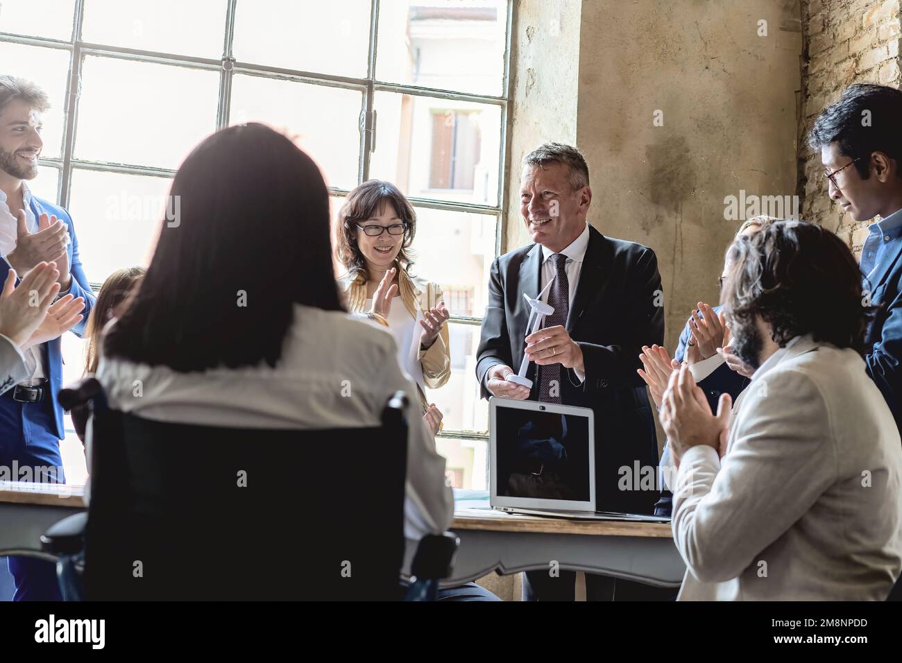 Eine vielfältige Gruppe von Menschen, die an einem Tisch sitzen und einem Mann applaudieren, der ein Modell einer Windturbine in einem Büro hält. Teamarbeit feiern Stockfoto