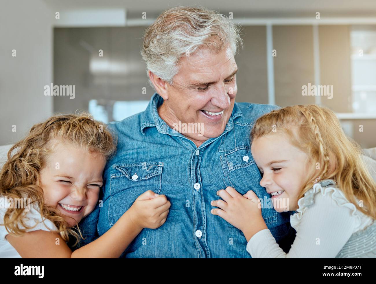 Eine glückliche dreiköpfige Familie, die sich auf dem Sofa entspannen kann. Lächelnder Opa, der zu Hause auf seine beiden Enkelinnen aufpasst Stockfoto
