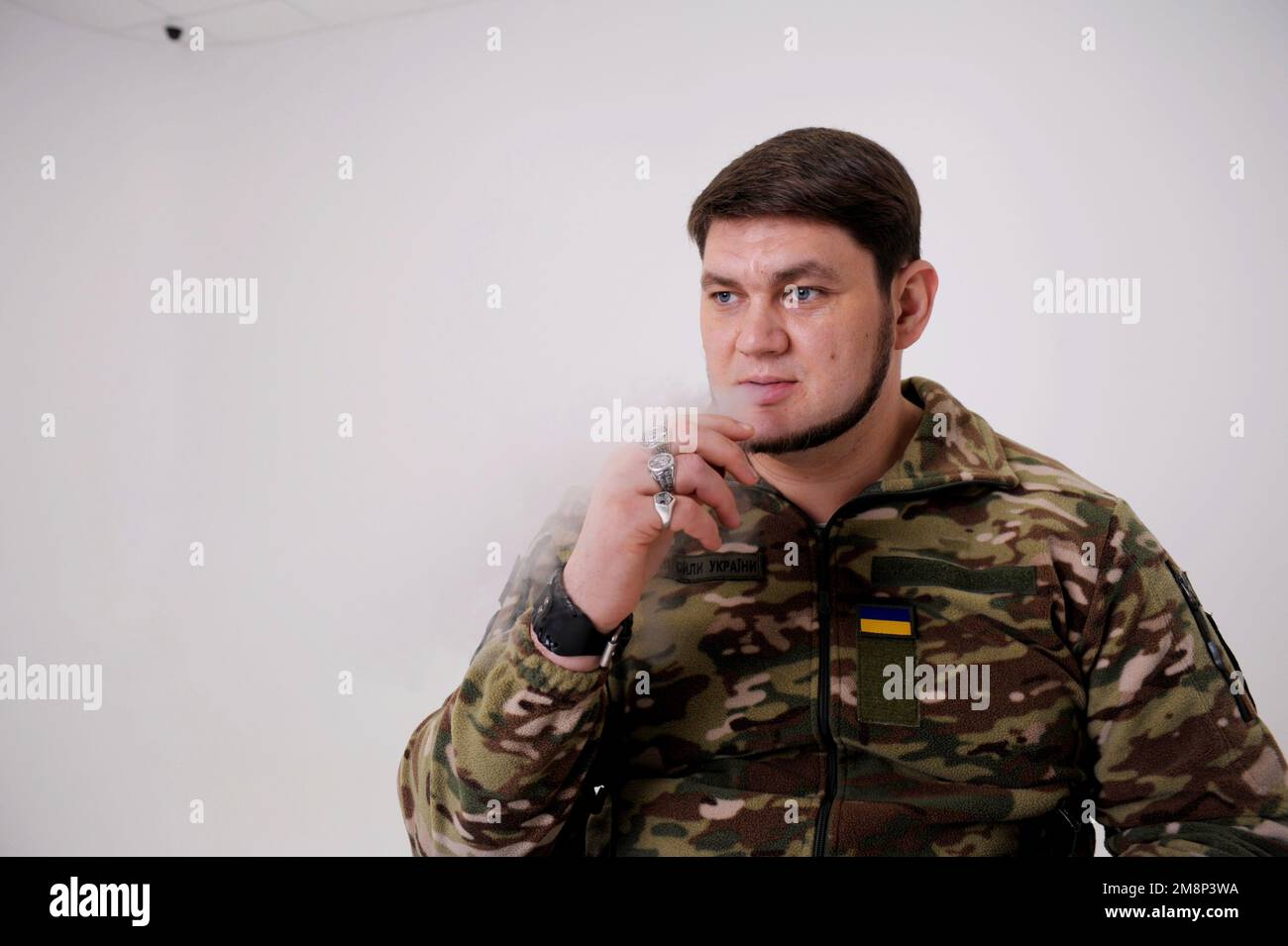 Militärmann in Uniform sitzt und raucht Er atmet Rauchpuppen aus Er hat Ringe an seinen Fingern Emblem der Ukraine die ukrainische Flagge ist auf Schutzjacke genäht Erwachsener attraktiver Mann 35 Jahre alt Stockfoto