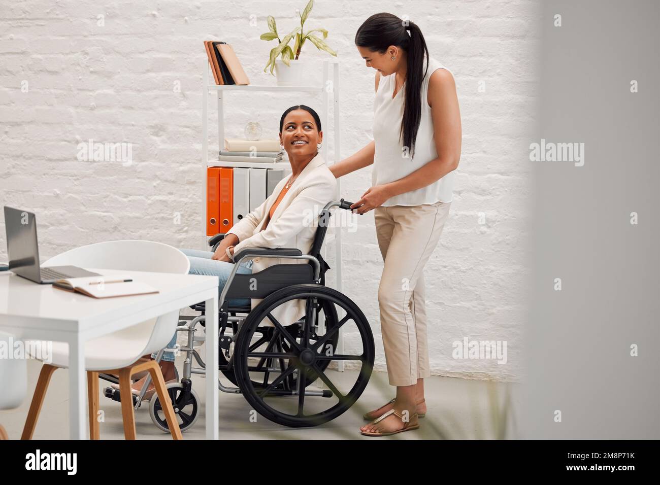 Junge gemischte Geschäftsfrau, die einer Kollegin im Rollstuhl bei der Arbeit in ihr Büro hilft. Hispanischer Geschäftsmann mit Behinderung, dem geholfen wird Stockfoto