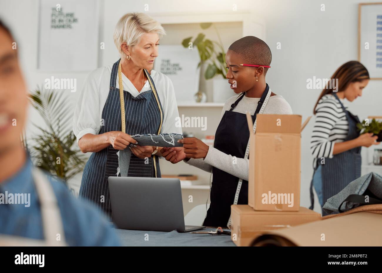Zwei Bekleidungsdesigner verwenden einen Laptop, während sie in einem Meeting mit Material arbeiten. Junge afroamerikanische Schneiderin, die während der Arbeit spricht Stockfoto