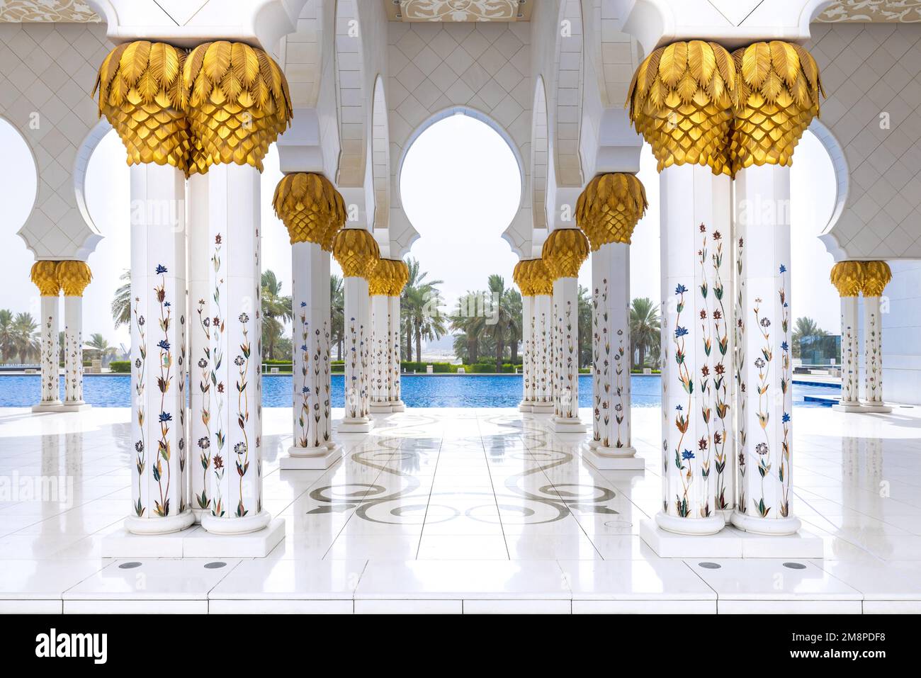 Eine weiße Marbe-Kolonnade mit goldenen Kapitellen führt zum Außenpool mit Palmen im Hintergrund der Scheich-Zayed-Moschee Stockfoto