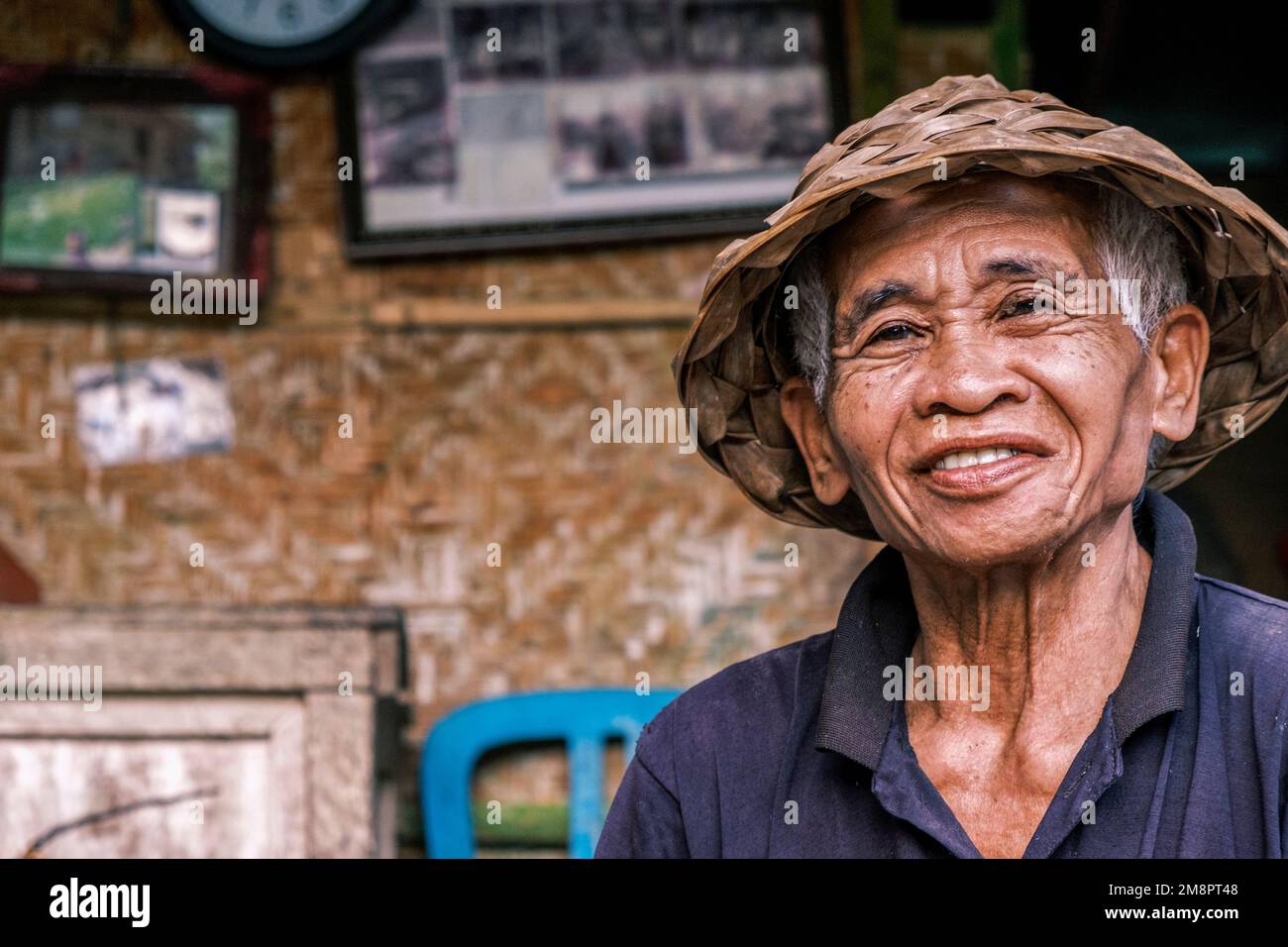 Horizontales Porträt eines südasiatischen Seifenmanns mit einem traditionellen balinesischen kegelförmigen Hut. Ein älterer Mann sieht lächelnd in die Kamera. Stockfoto