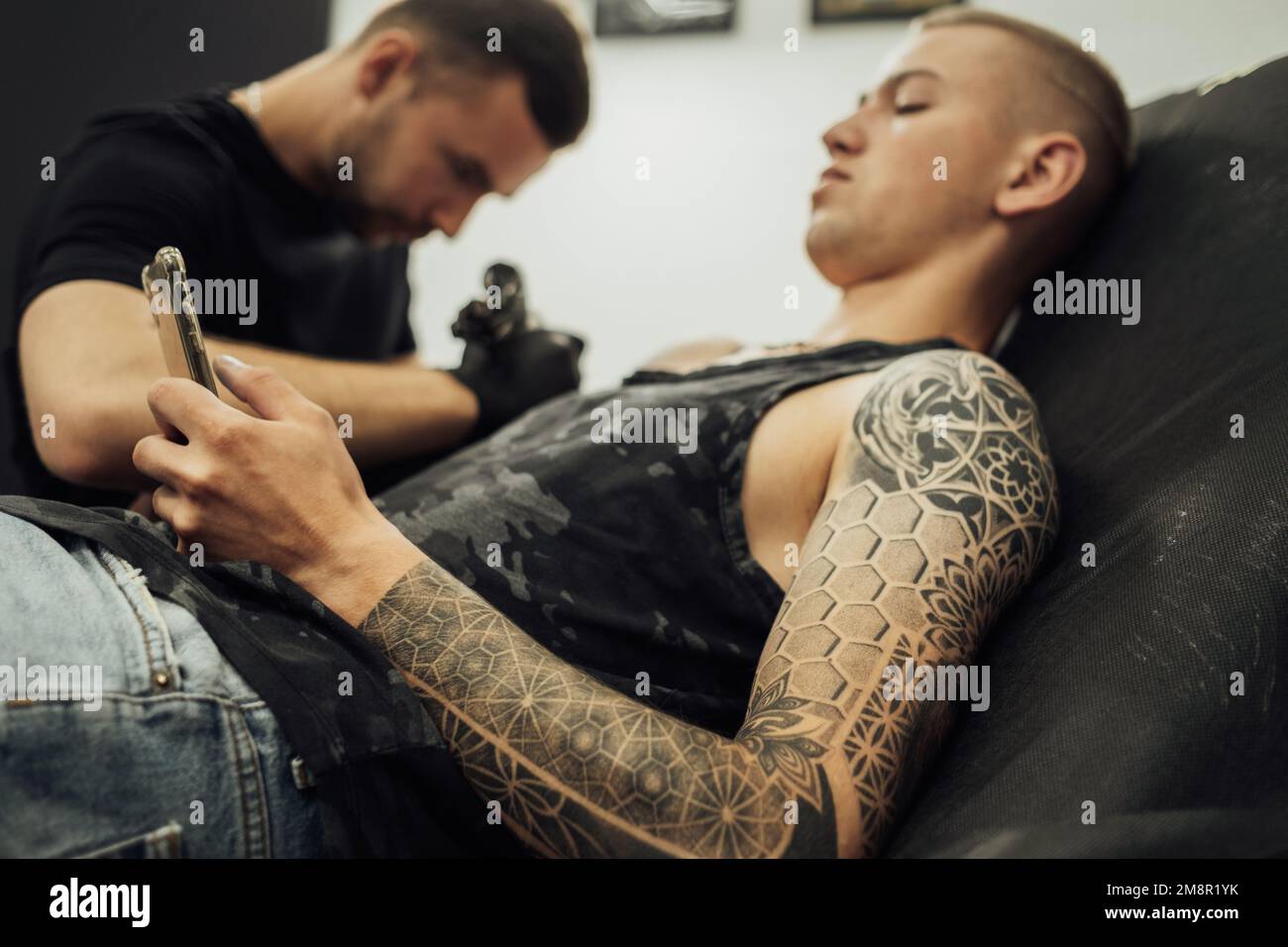 Tätowierter Mann, der während der Tattoo Session ein Smartphone benutzt, zeichnet männlicher Künstler auf die Haut des Kunden Stockfoto