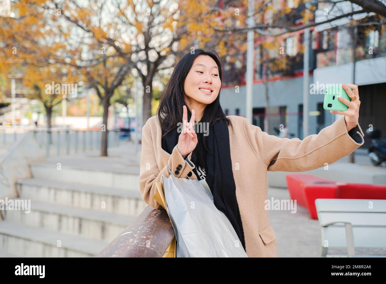 Fröhliche junge chinesin oder Asiatin, die lächelt und ein Selfie-Porträt macht, das Friedenszeichen auf dem Handy macht, nachdem sie eingekauft und über die sozialen Medien mit einer Smartphone-App geteilt hat. Hochwertiges Foto Stockfoto