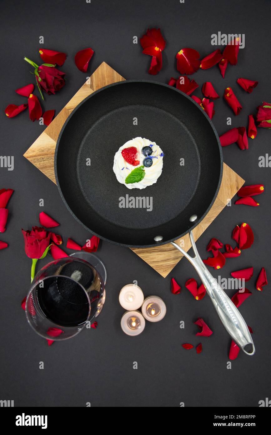 Valentinstag-Konzept mit roten Rosenblütenblättern auf schwarzem Hintergrund und einer Pfanne auf einem Holzbrett und einem pavlova-Kuchen in der Pfanne. Ein Glas Stockfoto