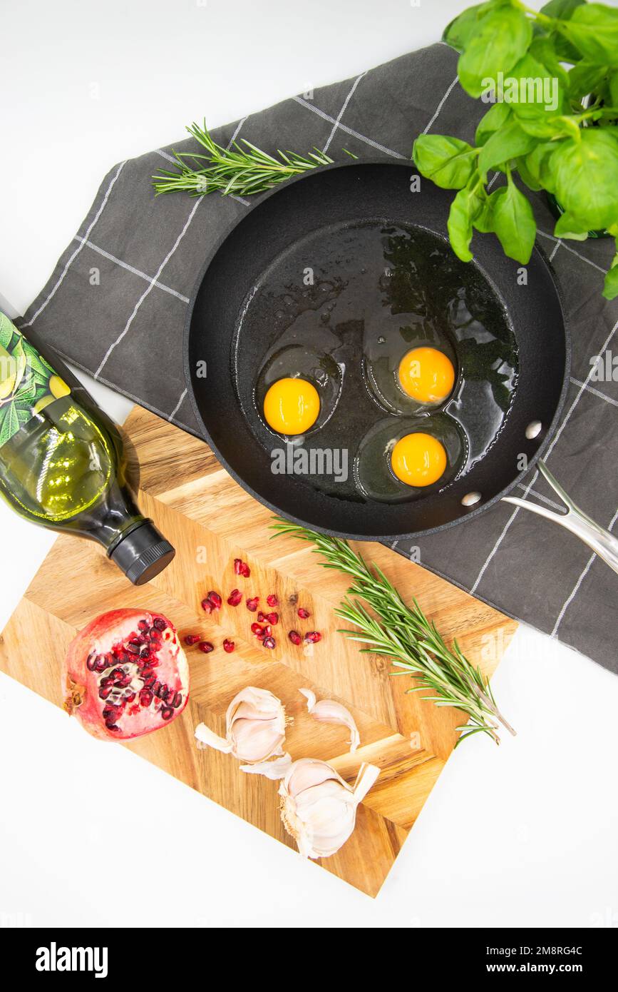 Schwarze Pfanne mit rohen Eiern auf einem dunklen karierten Handtuch neben einem hölzernen Kochbrett mit rotem Granatapfel und Knoblauch. Speisekonzept. Ansicht von bis Stockfoto