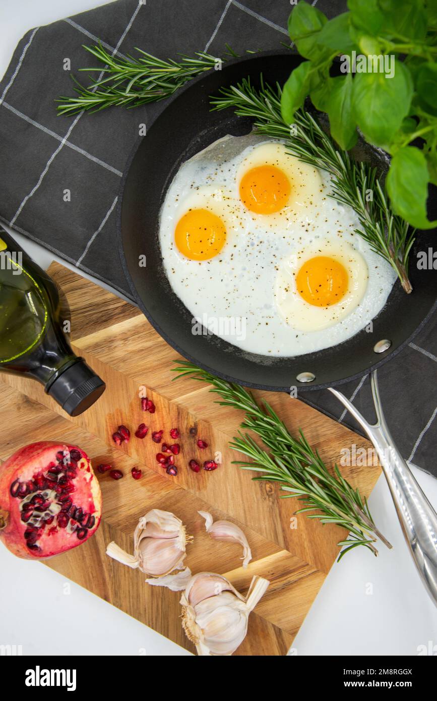 Frühstück oder Brunch, Spiegeleier in schwarzen Pfannen und roter Granatapfel mit Knoblauch und Basilikum. Ansicht von oben. Stockfoto
