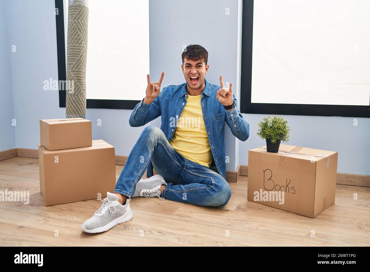 Junger lateinamerikanischer Mann, der im neuen Haus auf dem Boden sitzt und mit verrücktem Ausdruck schreit und mit erhobenen Händen ein Rocksymbol macht. Musikstar. Schweres Konzept. Stockfoto