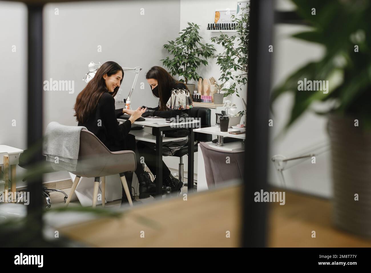 Nagelservice. Glückliche Kundin, die ihre Nägel im Schönheitssalon gemacht hat. Maniküre-Spezialist trägt Schutzmaske und Nägel für weibliche Klientin in Stockfoto