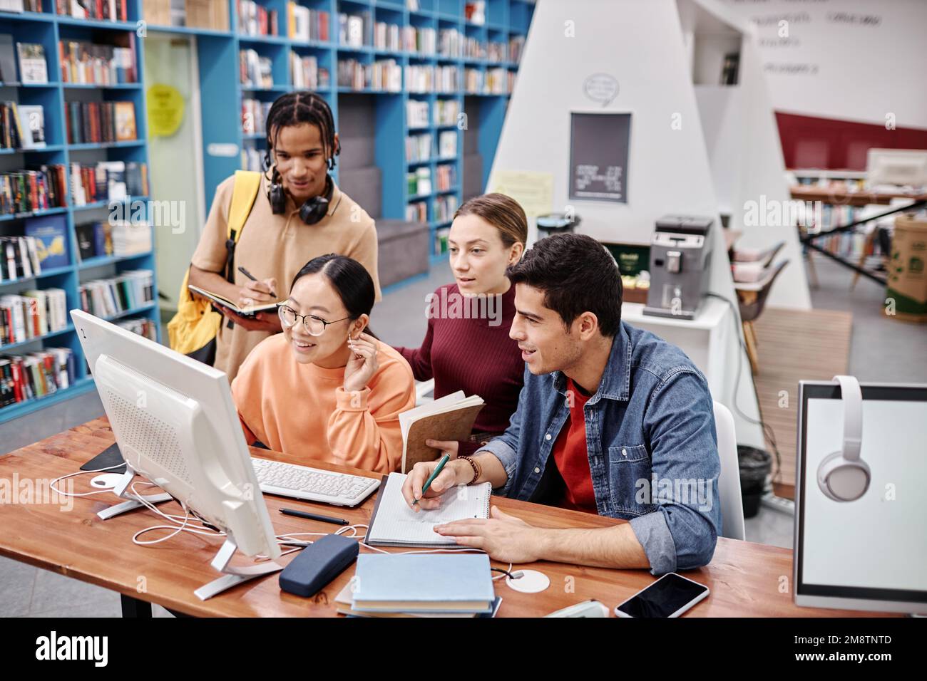 Vielfältige Gruppe von Studenten, die Computer zusammen in farbenfrohen Bibliotheksräumen nutzen und gerne lernen Stockfoto