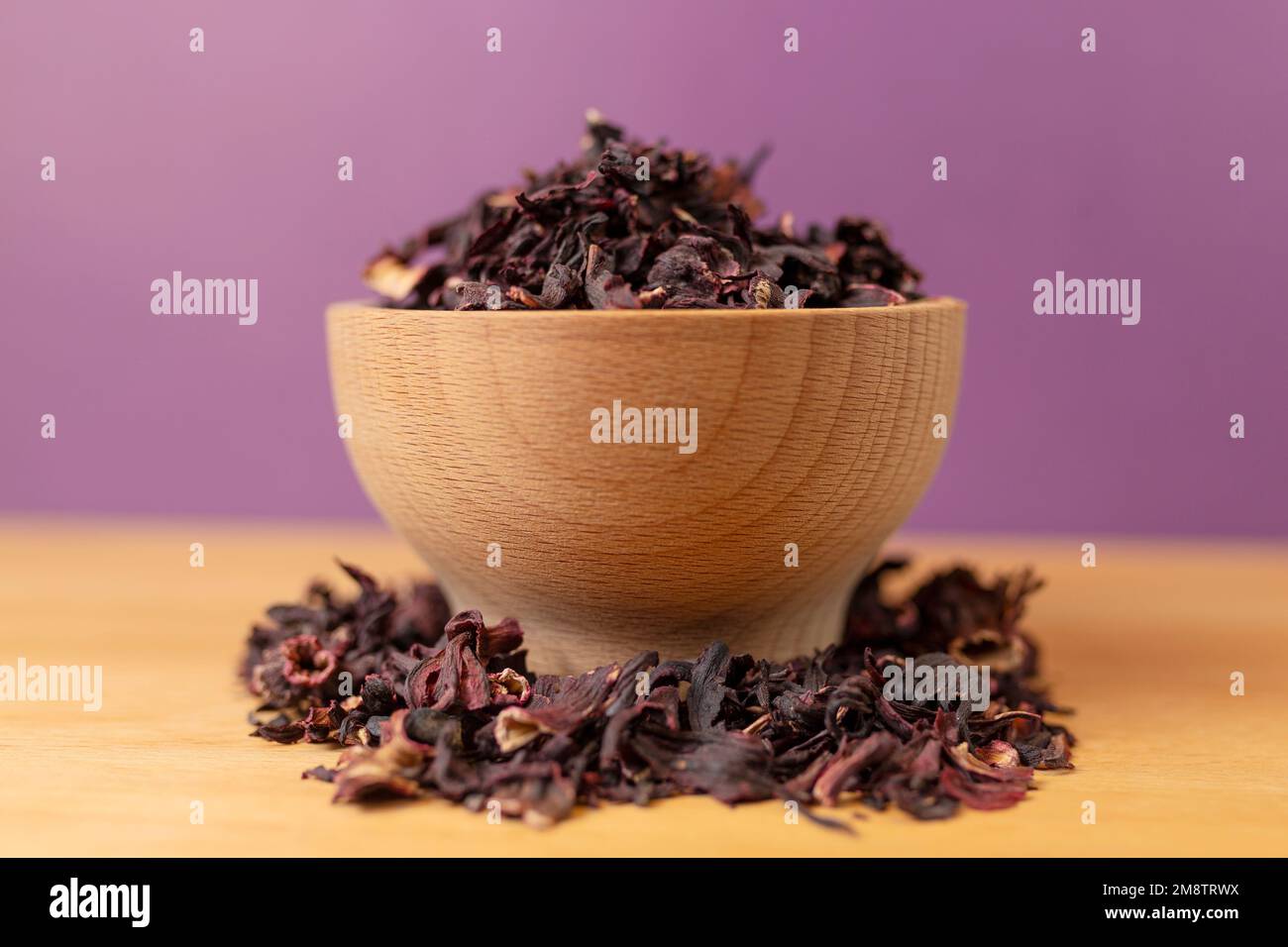 Trocknen Sie Hibiskus-Tee in der Schüssel auf Holzbrett. Violetter Hintergrund dahinter. Kräuter. Kräutertee. Lebensmittelzutat. Stockfoto