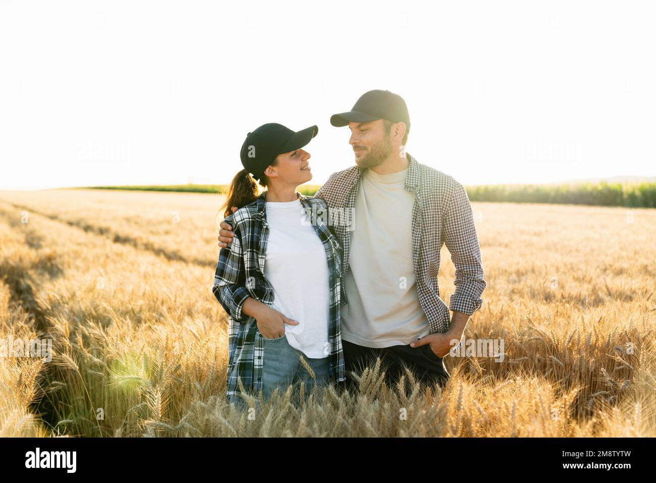 Ein paar Bauern in karierten Hemden und Kappen stehen bei Sonnenuntergang auf dem landwirtschaftlichen Weizenfeld umarmt Stockfoto