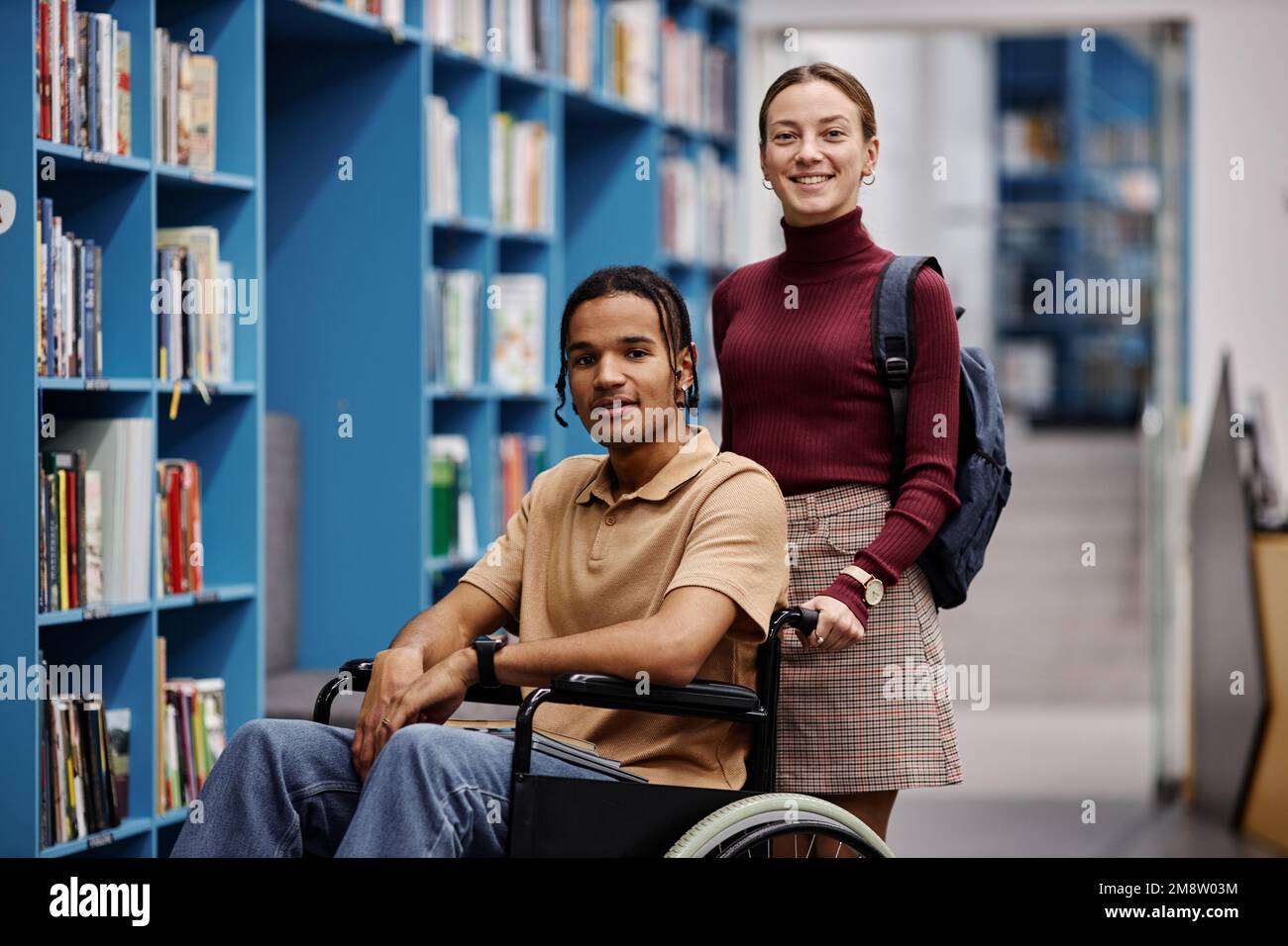 Porträt einer lächelnden jungen Frau, die einer behinderten Schülerin in der Bibliothek hilft und beide in die Kamera schaut Stockfoto