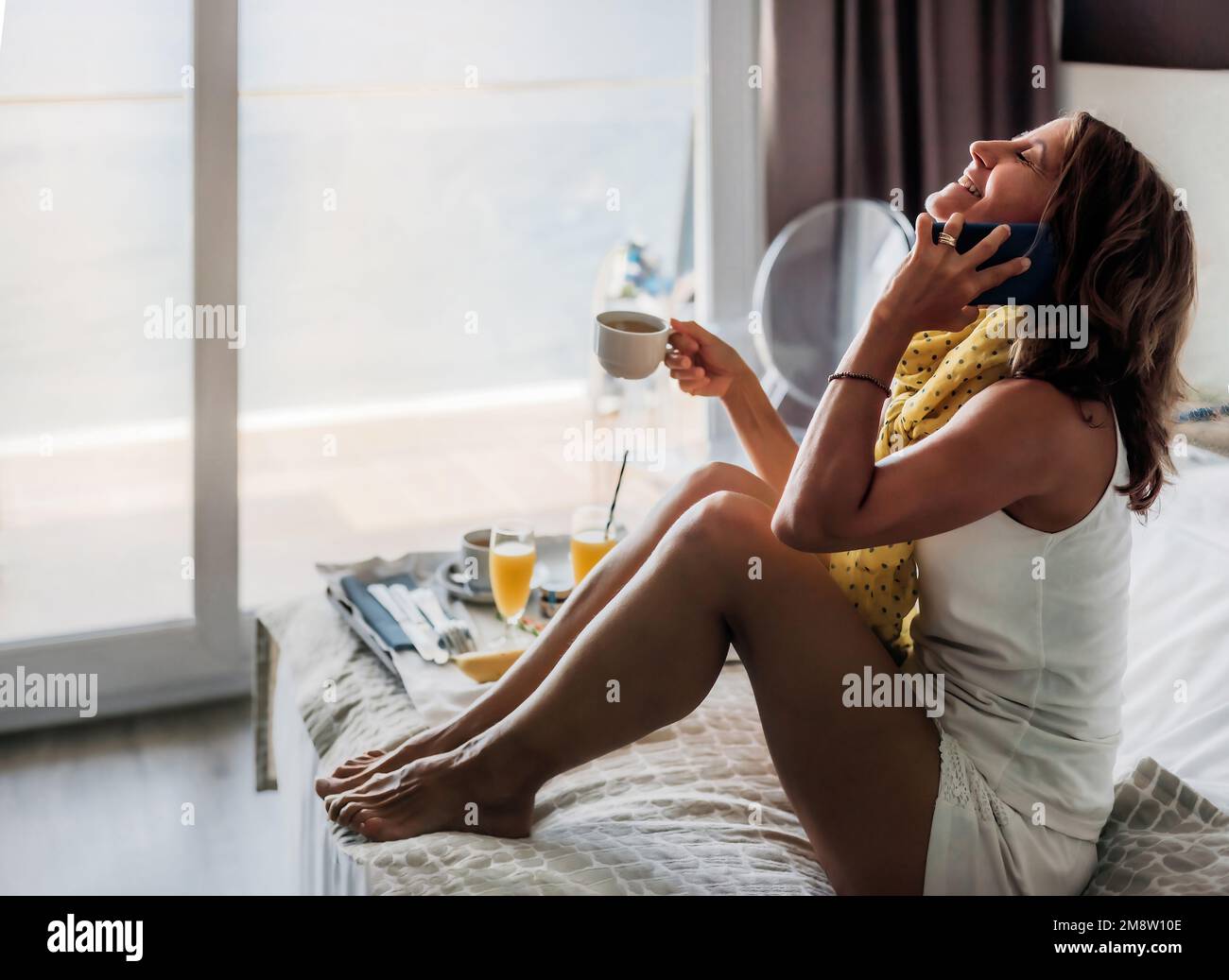 Eine weiße Frau mittleren Alters spricht am Telefon und lächelt, während sie auf einem Hotelbett frühstückt und eine Tasse Kaffee in der Hand hält. Tageslicht. Stockfoto