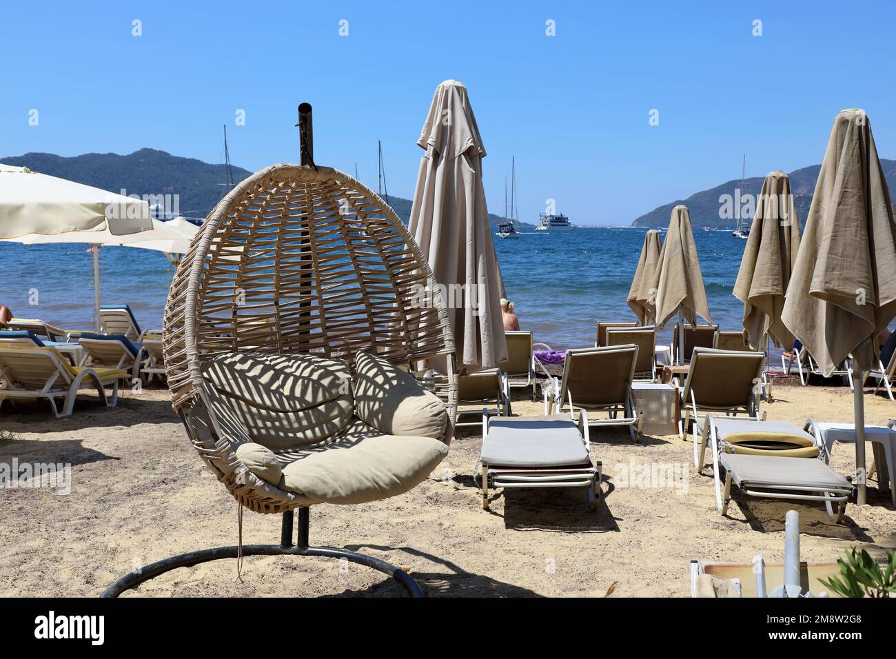 Korbstühle am Strand mit Sonnenschirmen und Liegestühlen. Sonniges Resort, Blick auf Berge und Yachten in der Ferne Stockfoto