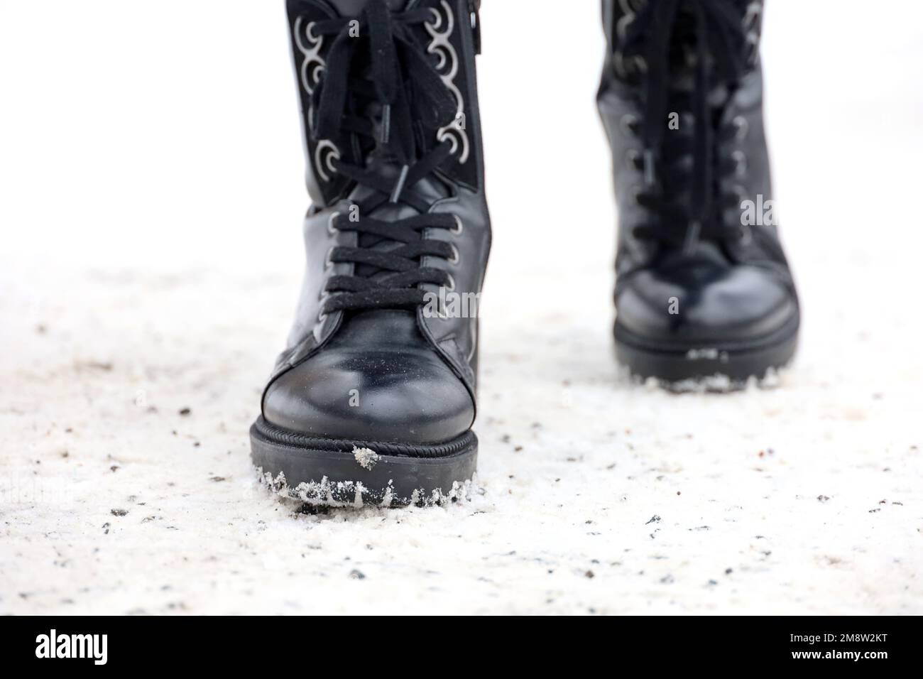 Weibliche Beine in schwarzen Lederstiefeln mit Schnürung auf einem Schnee, der mit einer Eisschutzmischung bedeckt ist. Frau, die auf der Winterstraße spaziert, warme Schuhe für kaltes Wetter Stockfoto