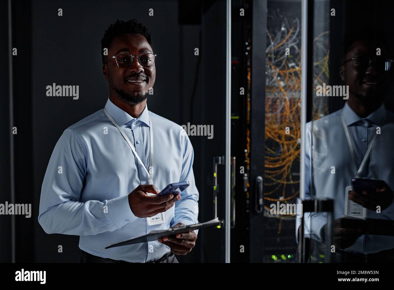 Porträt eines lächelnden Schwarzen als Netzwerktechniker, der im Rechenzentrum neben dem Serverschrank steht und in die Kamera und den Kopierbereich schaut Stockfoto