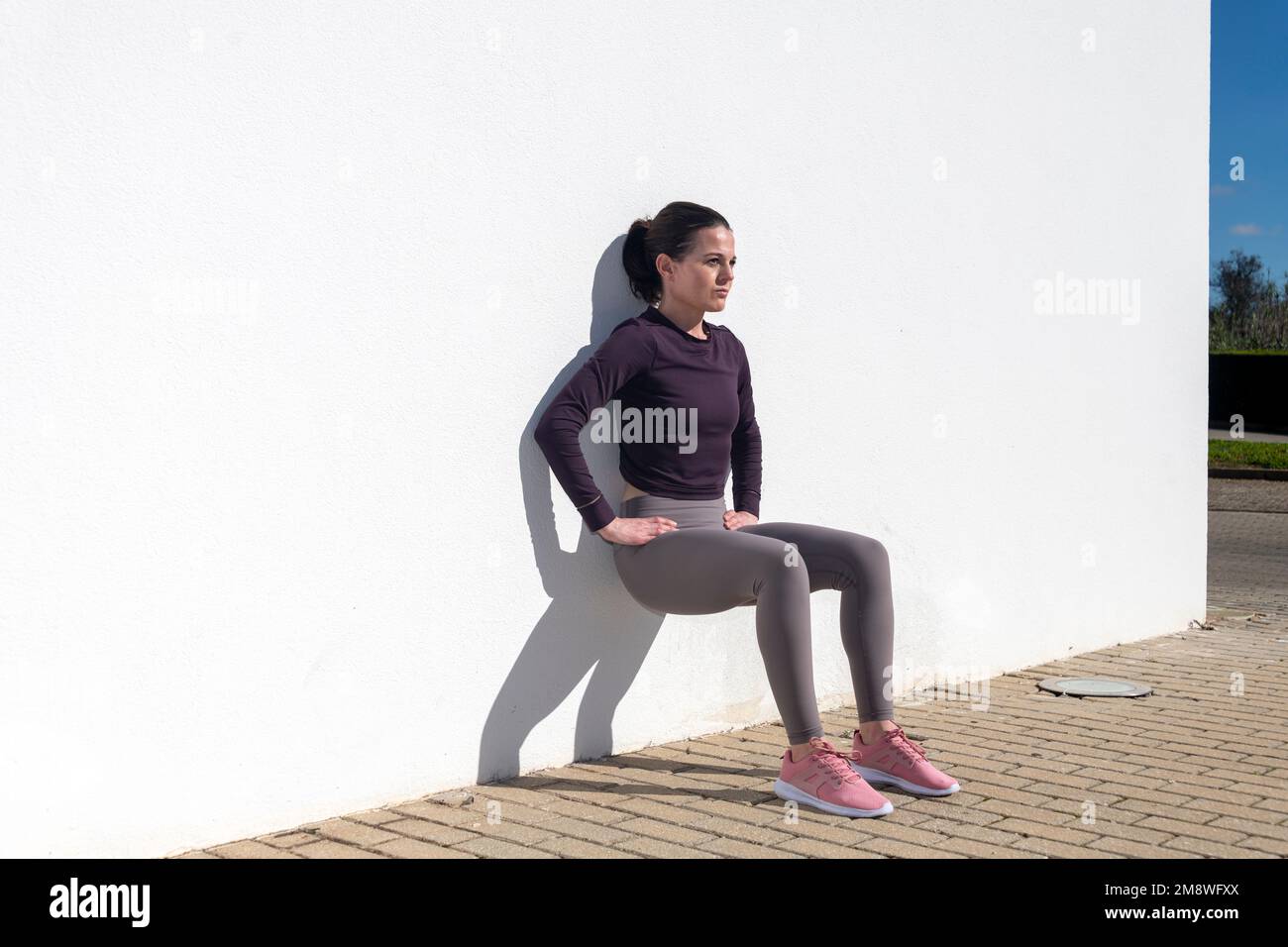 Sportliche Frau, die draußen in der Sonne Squats gegen eine weiße Wand macht. Stockfoto